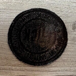 May include: A round, black patch with a textured surface. The patch has a circular design with text around the edge and a central design. The patch is on a wooden surface.