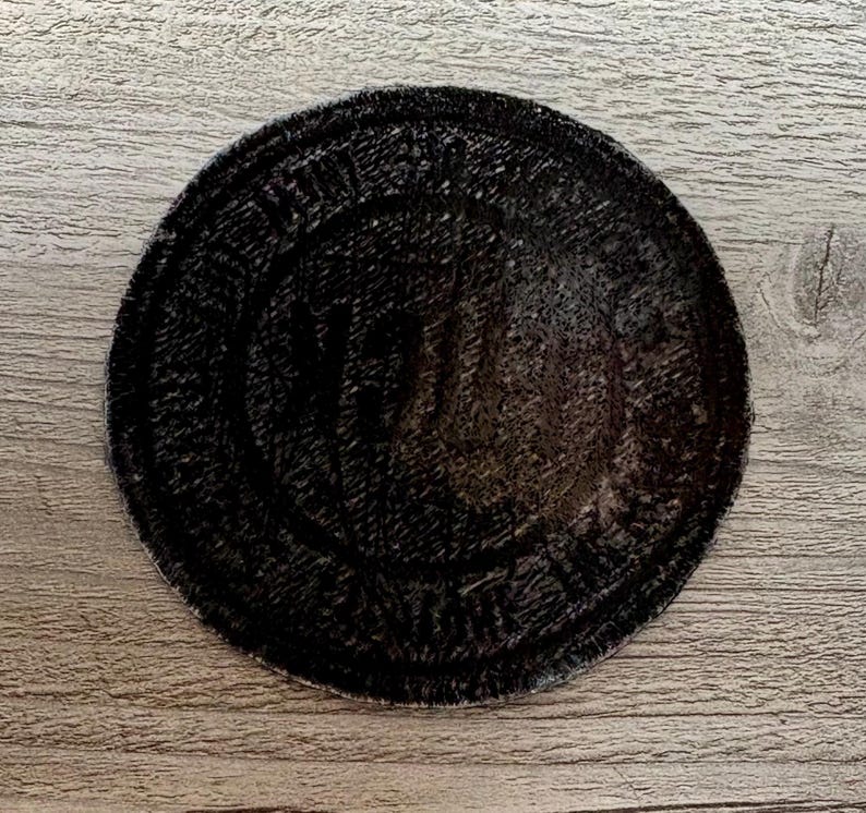 May include: A round, black patch with a textured surface. The patch has a circular design with text around the edge and a central design. The patch is on a wooden surface.