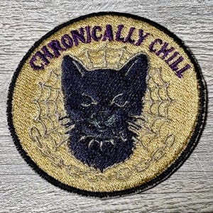 Può includere: Toppa ricamata con la faccia di un gatto nero e le parole "CHRONICALLY CHILL" in viola. Il gatto è centrato su uno sfondo dorato con un disegno a ragnatela. La toppa ha un bordo nero.
