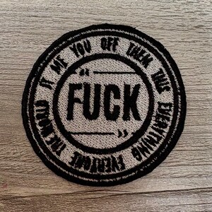 May include: Round, black and white embroidered patch with the word "FUCK" in large, bold letters in the center. The text "IT ME YOU OFF THEM THIS EVERYTHING" is in a circular pattern around the center. The patch is on a light wood surface.