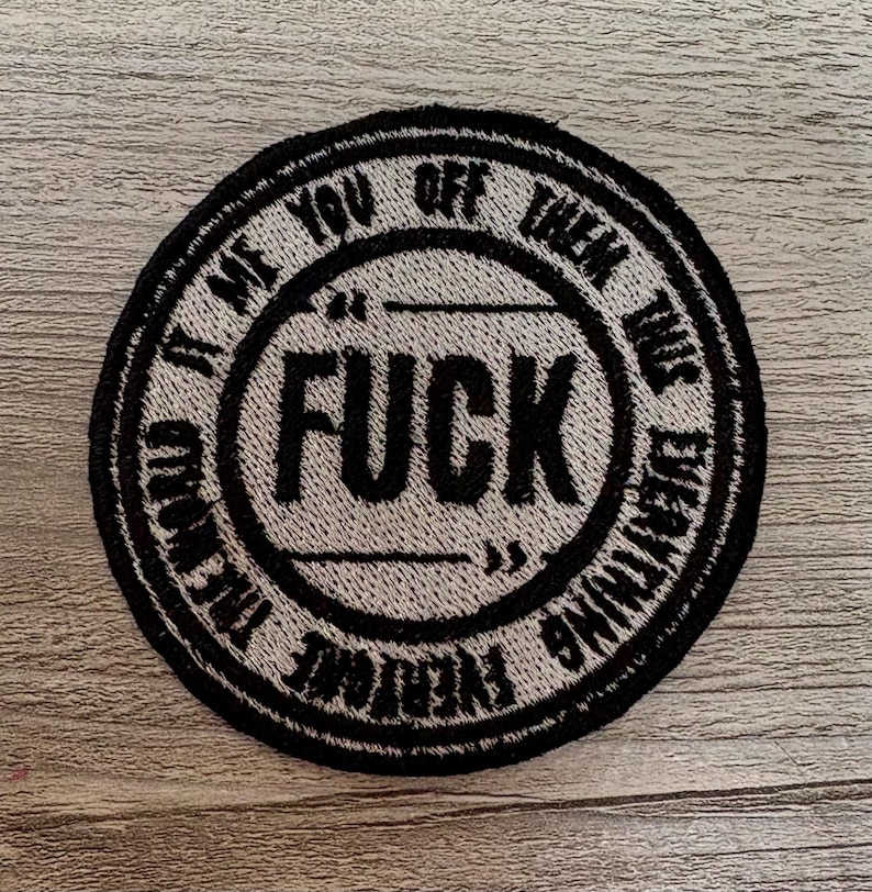 May include: Round, black and white embroidered patch with the word "FUCK" in large, bold letters in the center. The text "IT ME YOU OFF THEM THIS EVERYTHING" is in a circular pattern around the center. The patch is on a light wood surface.