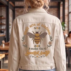 Puede incluir: Una chaqueta vaquera de color crema con un bordado en la espalda. El diseño muestra una abeja central rodeada por una corona floral y el texto "small acts of kindness make the Sweetest honey" en tonos marrón y amarillo.