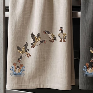 May include: Two rectangular kitchen towels, one in beige and one in dark gray, hanging side by side. Each towel features an embroidered sequence of geese in flight, with one goose diving into blue water, creating a splash effect at the bottom.