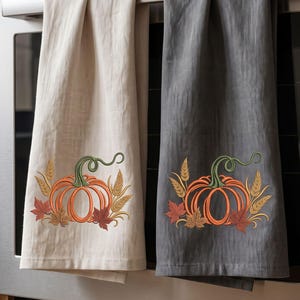 May include: Two kitchen towels hanging on an oven handle. One towel is cream-colored and the other is dark gray. Both feature an embroidered design of an orange pumpkin with a green stem, surrounded by golden wheat stalks and brown autumn leaves.