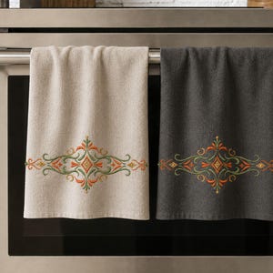 May include: Two rectangular kitchen towels hanging on an oven handle. One towel is light beige and the other is dark charcoal gray. Both feature an identical embroidered design in the center with orange and green scrolling floral patterns.