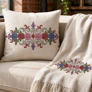 May include: A square beige throw pillow and a matching beige throw blanket with fringe, both featuring an identical embroidered design of blue, red, and pink roses arranged in a symmetrical floral pattern with green leaves and purple accents.