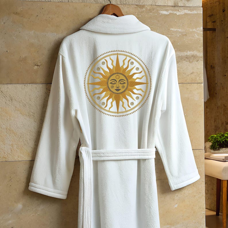 May include: White terry cloth robe with a gold sun design embroidered on the back. The sun has a smiling face and radiating lines. The robe has a shawl collar, long sleeves, and a matching belt. The robe is hanging on a wooden hanger.