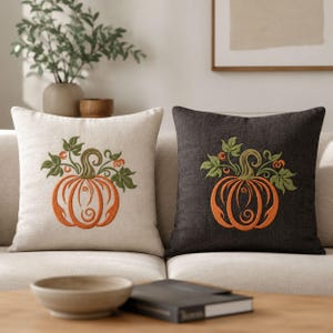 Pode incluir: Duas almofadas quadradas, uma em bege claro e outra em cinza carvão escuro, cada uma com um design bordado central de uma abóbora laranja com folhas e vinhas verdes. O tecido apresenta uma textura tecida visível em ambas.