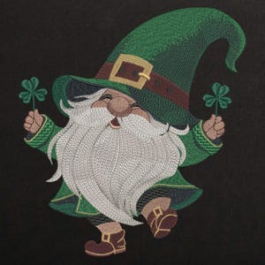 May include: Embroidered image of a cheerful leprechaun wearing a green hat with a brown belt and buckle, green coat, and brown shoes. The leprechaun has a long white beard and is holding two green shamrocks against a black background.