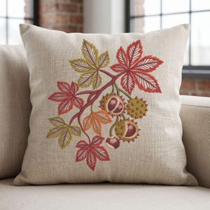 May include: A square beige throw pillow featuring an embroidered design of a chestnut branch with green and red leaves, along with several spiky green chestnut husks and smooth brown chestnuts centered on the front of the fabric cover.