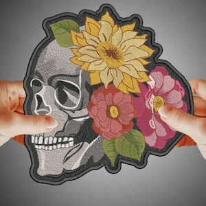 Könnte beinhalten: Gestickter Aufnäher mit einem grauen Schädel und einem floralen Design. Die Blumen sind gelb, rosa und grün.