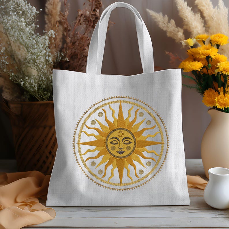 May include: White tote bag featuring a golden sun design with a smiling face and radiating beams. The sun is encircled by a decorative border. The bag has two white handles and is made of a light-colored fabric. The bag is sitting on a wooden surface.