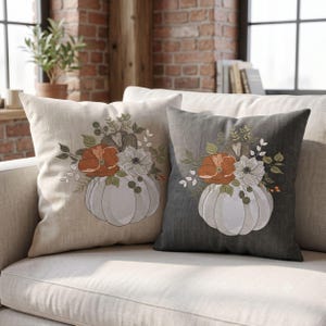 May include: Two square throw pillows featuring embroidered designs of white pumpkins topped with orange and white flowers and green foliage. One pillow is light beige, and the other is dark charcoal grey, both showcasing detailed stitching.