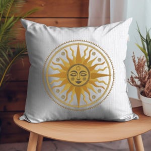 May include: White square throw pillow featuring a golden sun design with a smiling face. The sun is surrounded by a golden circle with decorative elements. The pillow is placed on a wooden chair, with plants in the background.