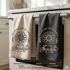 Könnte beinhalten: Zwei Küchentücher, eines beige und eines dunkelgrau, hängen an einer Metallstange. Jedes Tuch zeigt ein Sonnenblumenmuster mit dem Text "Wenn du die Sonne nicht finden kannst, sei die Sonne."