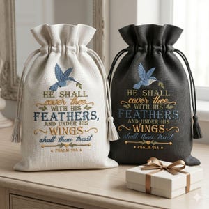 May include: Two drawstring bags, one cream and one charcoal gray, each featuring an embroidered blue hummingbird and the text: He shall cover thee with his feathers, and under his wings shalt thou trust. Psalm 91:4. A small white gift box sits in front.