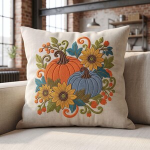 May include: A square throw cushion with a cream-coloured fabric cover featuring an embroidered design of an orange pumpkin, a blue pumpkin, yellow sunflowers, green leaves, and orange berries arranged in a central decorative cluster.