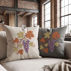 May include: Two square throw pillows, one in light beige and one in dark gray, each featuring an embroidered design of a grape cluster with green and orange leaves. The embroidery displays detailed vine tendrils and purple grapes on both cushions.