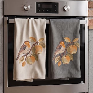 May include: Two kitchen towels hanging on an oven handle. One towel is cream-colored and the other is dark gray. Both feature an identical embroidered design of a small bird perched on a branch with two round fruits and several leaves in shades of orange, brown, and gold.