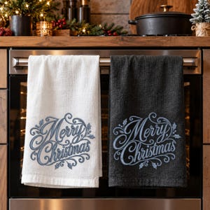 Può includere: Un canovaccio bianco e uno grigio scuro appesi alla maniglia di un forno. Ogni asciugamano presenta un ricamo blu con la scritta Merry Christmas circondata da ghirigori decorativi e foglie di agrifoglio per un tocco festivo.