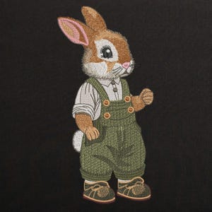 May include: Embroidered patch of a brown bunny wearing green overalls and a white shirt with a button. The bunny has brown ears, a white face, and black eyes.