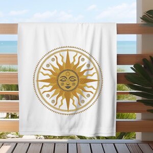 May include: White beach towel featuring a detailed sun design with a smiling face, radiating golden rays, and a decorative border. The sun design is centered on the towel, which is hanging on a wooden railing with a view of the ocean.