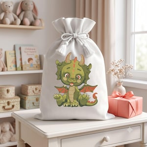 Könnte beinhalten: Ein weißer Geschenksack aus Stoff mit Kordelzug, verziert mit einer Cartoon-Illustration eines grünen Drachen mit orangefarbenen Flügeln und Hörnern. Der Sack hat einen gerafften Verschluss mit weißen Kordeln und Quasten.