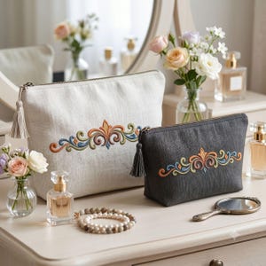 May include: A large beige linen pouch and a smaller charcoal grey linen pouch, both featuring a central colourful embroidered scroll design and a matching tassel on the zipper pull. The pouches are arranged with perfume bottles and jewellery.