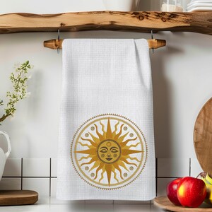 May include: White kitchen towel with a waffle weave texture, featuring an embroidered sun design in shades of gold. The sun has a smiling face and is surrounded by a decorative border. The towel is hanging on a wooden rack.