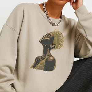 Può includere: Felpa beige con un'illustrazione ricamata di una donna che guarda in alto. L'illustrazione è nei toni del nero, oro e beige. La donna indossa un copricapo e un top dorato. È visibile anche una collana a catena d'argento.