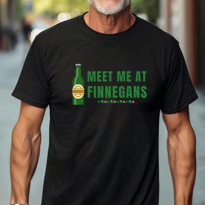 Puede incluir: Una camiseta negra con un gráfico de una botella de cerveza verde y el texto "Meet me at Finnegans".