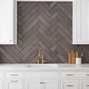 May include: A modern kitchen with white cabinets, a white countertop, and a dark wood herringbone backsplash. A gold faucet and hardware complement the design. Includes a cutting board, utensils, and a gas stove.