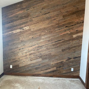 May include: A room with a wall covered in reclaimed wood planks. The planks are arranged in a diagonal pattern and are a mix of brown, gray, and black colors. The wall is accented with a white electrical outlet. The floor is a light gray concrete.