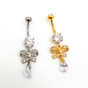 May include: Two belly button rings with a bow and a teardrop-shaped crystal. One ring is silver with clear crystals and the other is gold with clear crystals.