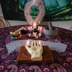 May include: A table setting with a purple and black paisley tablecloth. There are candles, a mortar and pestle, a crystal, a singing bowl, and a hand with lines on it.