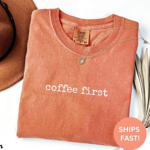 May include: A coral-colored Comfort Colors t-shirt with the phrase "coffee first" in white lettering. A gold necklace with a pendant rests on the shirt. A brown hat is visible in the background.