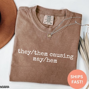 May include: A brown t-shirt with the text "they/them causing may/hem" printed in white. The shirt is made of a soft, comfortable material.