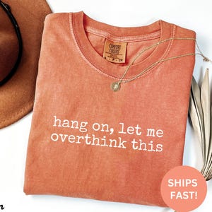 May include: A burnt orange t-shirt with the text "hang on, let me overthink this" printed in white.