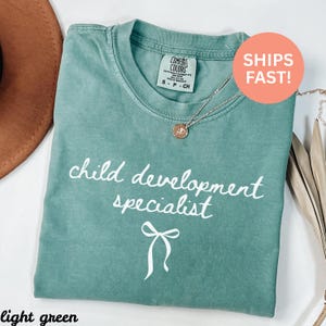 May include: A light green t-shirt with the words "child development specialist" in white script. A bow graphic is below the text. The shirt has a crew neck and short sleeves. A gold necklace with a pendant is on the shirt.