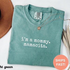 Puede incluir: Camiseta verde claro con el texto "i'm a mommy. mamacita." en blanco. Un collar dorado con un colgante est&aacute; drapeado sobre la camiseta. La camiseta est&aacute; doblada y tiene una etiqueta Comfort Colors.