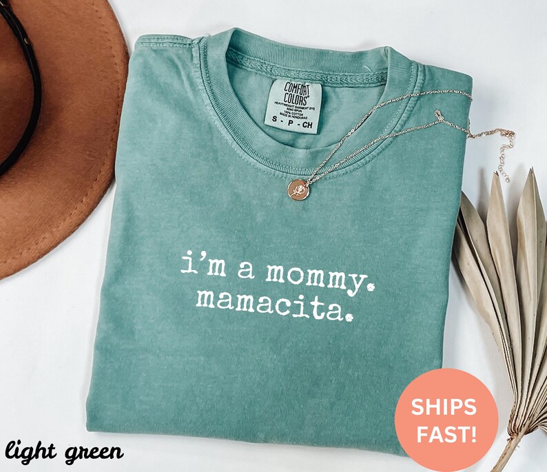 Puede incluir: Camiseta verde claro con el texto "i'm a mommy. mamacita." en blanco. Un collar dorado con un colgante est&aacute; drapeado sobre la camiseta. La camiseta est&aacute; doblada y tiene una etiqueta Comfort Colors.