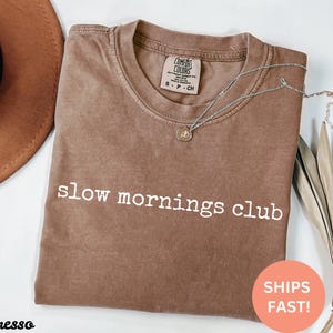 May include: A brown t-shirt with the text "slow mornings club" printed in white. The shirt is made of a soft, comfortable material and has a relaxed fit.