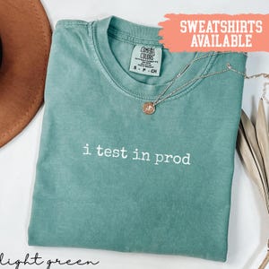 Puede incluir: Camiseta verde claro con el texto "i test in prod" en blanco. Un collar dorado con un colgante descansa sobre la camiseta. La camiseta tiene cuello redondo y mangas cortas. La etiqueta dice "Comfort Colors".