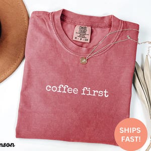 May include: Crimson-colored t-shirt with the phrase "coffee first" in white lettering. A gold necklace with a pendant rests on the shirt. The shirt has a Comfort Colors tag. A brown hat and dried leaves are in the background.