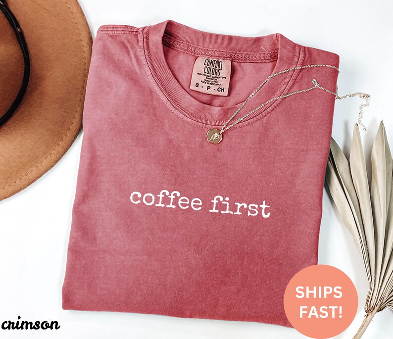 May include: Crimson-colored t-shirt with the phrase "coffee first" in white lettering. A gold necklace with a pendant rests on the shirt. The shirt has a Comfort Colors tag. A brown hat and dried leaves are in the background.