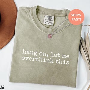 May include: A khaki green t-shirt with the text "hang on, let me overthink this" printed in white. The shirt is made of a soft, comfortable material.