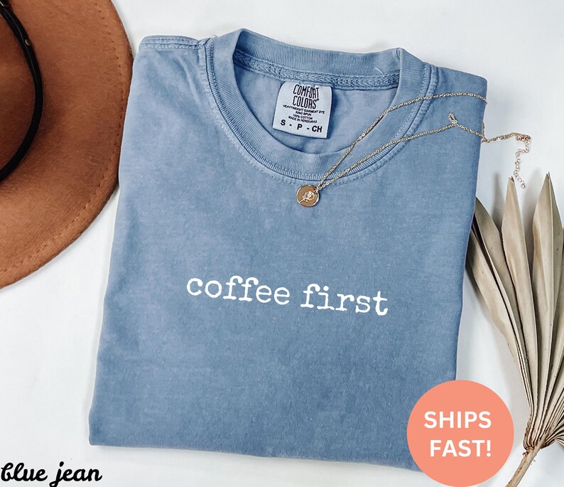 May include: A blue jean Comfort Colors t-shirt with the phrase "coffee first" in white lettering. A gold necklace with a pendant rests on the shirt. A brown hat and dried plant are in the background.