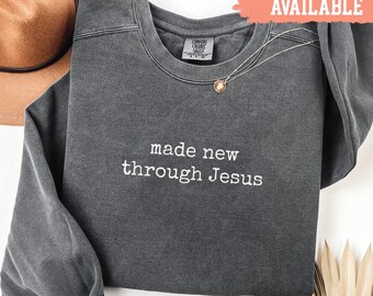 May include: A charcoal grey sweatshirt with the embroidered text "made new through Jesus". A gold pendant necklace rests on the shirt. A coral banner in the top right corner reads "SHIRTS AVAILABLE".