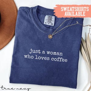 Può includere: T-shirt blu navy con la scritta bianca "just a woman who loves coffee". Una collana con pendente dorato è appoggiata sulla maglietta. Le parole "SWEATSHIRTS AVAILABLE" sono in uno striscione color pesca.