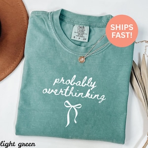 May include: Light green t-shirt with the phrase "probably overthinking" in white script and a bow graphic. The shirt has a crew neck and short sleeves. A gold necklace with a pendant is draped on the shirt.
